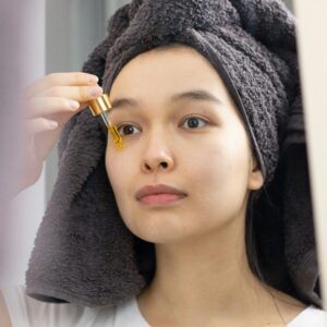 An Asian woman applies skincare serum with a dropper in front of a mirror.